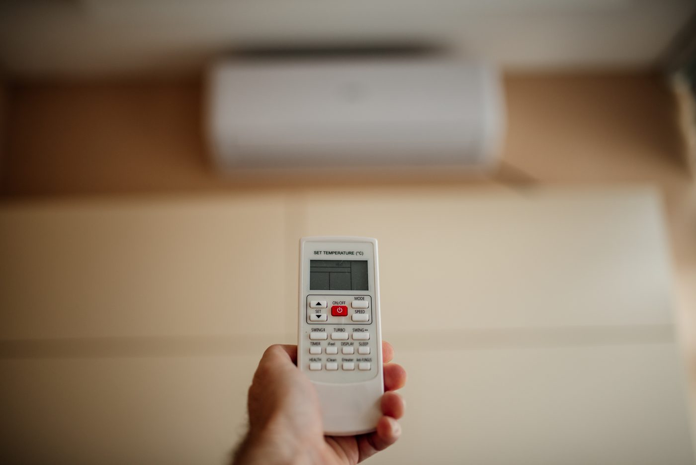 A hand holds an air conditioner remote control, pointing it at a wall-mounted indoor unit.